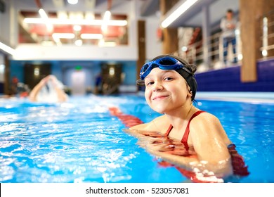 Girl Child Swimmer In A Red Bathing Suit On A Background Of The Pool Inside The Room With Glasses And A Cap.