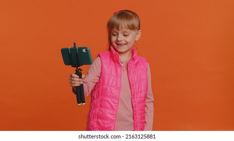 Girl Child Toddler Blogger Taking Selfie On Mobile Phone Selfie Stick, Communicating Video Call Online With Subscribers. Young Little Children Kid Teen Isolated Alone On Orange Studio Wall Background