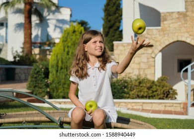 Girl Child Tossing, Playing, Juggling Green Apples, Sitting On Outdoor Chair On Green Lawn, Sunny Summer Day. Healthy Fresh Vitamin Food