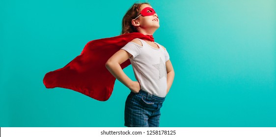 Girl Child Wearing Red  Costume Standing With Her Hands On Hips In Studio. Girl In Red Cape And Mask Looking Away On Blue Background.