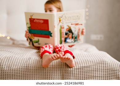 A girl in a Christmas pajama is reading a large book on the bed, focusing on her feet.