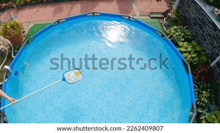 Girl cleaning leaves from surface of pool water with net skimmer. Top view, wide angle. Beautiful flowers grow around the pool. Maintenance of a frame pool with net skimmer