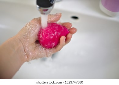 girl cleans in her hand a pink rosa beauty blender for make-up with water, on the background there is a bright bathroom and a wash basin