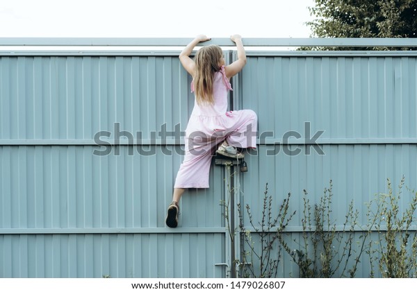 Girl climbing metal fence outdoor. Curious child\
on high white painted gates. Naughty kid playing outside, breaking\
rules. Childhood and youth concept. Restless teen entering private\
property