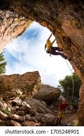 The Girl Climbs The Rock In The Shape Of An Arch, A Man Is Belaying A Climbing Partner,  Friends Train On Natural Terrain, The Well Coordinated Team. Fitness And Active Lifestyle.