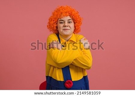 a girl in a clown costume and a bright wig hugs herself with her hands, studio portrait on a colored background