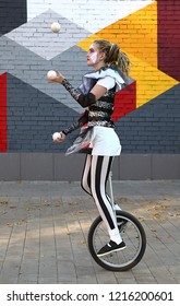 Girl clown rides a unicycle and juggles balls  in front of colorful wall outdoors