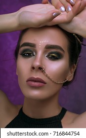 Girl with cool fashionable make-up posing in studio,matte lips,smokey gold eyes, beautiful make up and piercing chain from nose over purple dark background with magic glow. Holiday disco celebration
