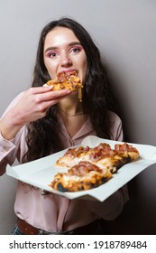 Girl Courier Eating Pinsa Pizza Romana Gourmet Italian Cuisine On Grey Background. Holding Scrocchiarella Traditional Dish. Food Delivery From Pizzeria. Pinsa With Meat, Arugula, Olives, Cheese.