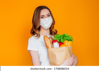 Girl Courier Volunteer Wearing A Medical Face Mask Holds A Paper Bag With Products, Vegan Vegetables, Herbs Isolated Over Orange Background, Quarantine, Coronavirus, Safe Food Delivery, Donation