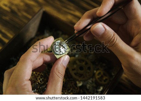 Girl craftsman dismantles clock. Repairing vintage watches. Female hands holding tweezers. Disassembly of clock on background of wooden table. Beautiful female fingers with pincers at work.