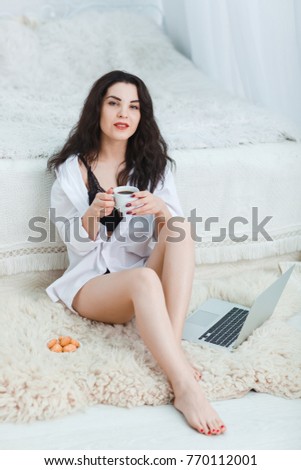 A girl with a cup of coffee in hands with a hot look sits on a rug by the bed in front of a white background
