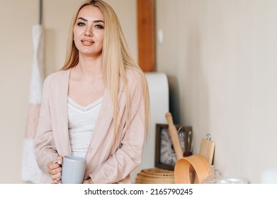 Girl With Cup In Home Clothes Is Standing In Kitchen. Close-up Portrait Of Young Mistress. Warm Processing. Good Morning Concept.