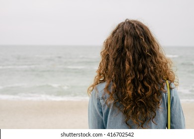 Girl With Curly Hair In A Denim Shirt Looking At Sea, View From The Back