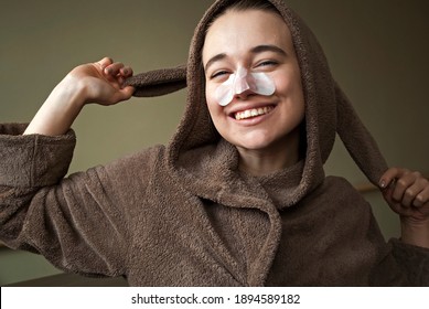 

Girl In A Cute Bathrobe, Dressing Gown With Bunny Ears. Woman Has Fun With A Facial Mask.A Woman Applies Cleansing, Moisturizing Strips To Her Nose,  Mask From Blackheads.