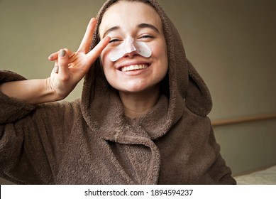 

Girl In A Cute Bathrobe, Dressing Gown With Bunny Ears. Woman Has Fun With A Facial Mask.A Woman Applies Cleansing, Moisturizing Strips To Her Nose,  Mask From Blackheads.