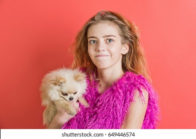 Girl Or Cute Woman With Natural Blond, Long, Wavy Hair Smiling With Closed Mouth On Young Face, No Makeup In Pink Feather Boa And Small, Spitz Dog, Pet In Hands On Red Wall. Fashion, Friendship