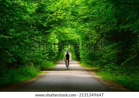 A girl cyclist is riding her road bike on the road in a beautiful thick green forest. Bikegirl in mystery forest. Whire roadbike. Black cycling suit.