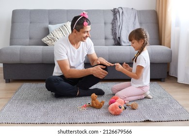 Girl and dad with sitting on floor near sofa and making manicure, daughter paints her father nails with nail polish, doing beauty procedures at home, funny pastime.