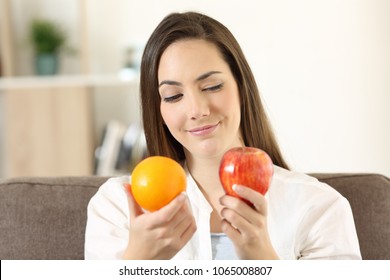 Girl deciding between two fruits an apple and an orange sitting on a couch in the living room at home