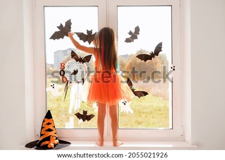 The girl decorates the window in the room with bats and Halloween ghosts standing on the windowsill. Preparing for a masquerade party. A girl in an orange witch dress.
