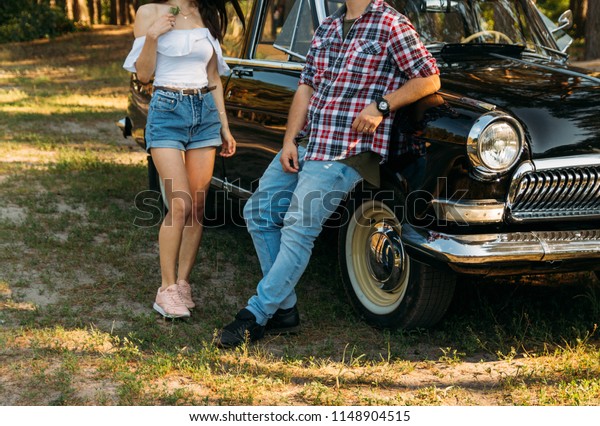 a girl\
in denim shorts and a white blouse, with a flower in her hands,\
shyly approaches a man in jeans and a red shirt in a cage stands on\
a vintage, retro car for dating.\
summer