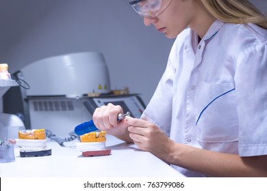 Girl Dental Technician In The Laboratory, In A Medical Dressing Gown, Clothes, Wearing Goggles, She Makes Dental Prosthesis, Cermet