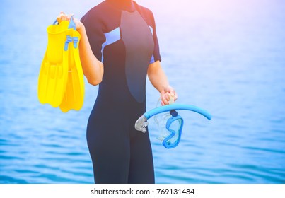 A Girl Diver In A Wet Suit Holds A Mask With A Blue Tube In Her Hands And A Yellow Fin In The Other Hand.