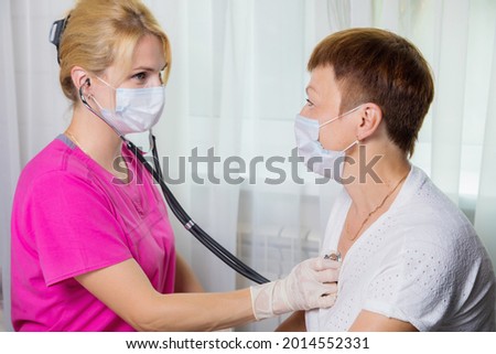A girl doctor in a mask listens with a stethoscope to the chest of an adult woman. A woman in the doctor's office complains of a cough.