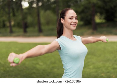 Girl doing exercises with dumbbells in the park. She smiles