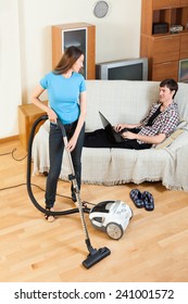 Girl Doing Room Cleaning With Vaccuum Cleaner While Man  With Laptop  Resting Over Sofa