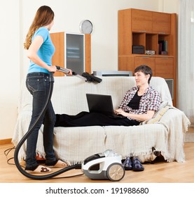 Girl Doing Sofa Cleaning With Vaccuum Cleaner While Man Resting Over Sofa