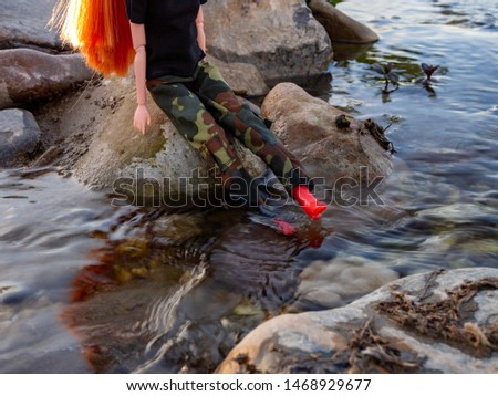Girl doll with red hair, green army pants, black T-shirt and red boots sitting on a rock next to a stream. Feet in the water. 
