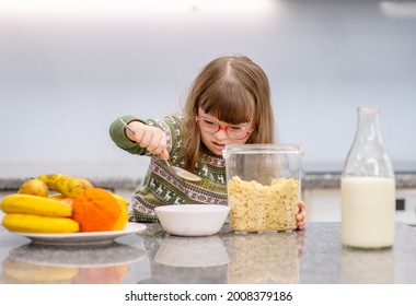 A Girl With Down Syndrome Is Having Breakfast With Cereals And Milk At Home In The Kitchen. Ordinary Childcare In A Family For Children With Disabilities