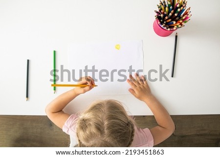 The girl draws with colored pencils sitting at a white table. Top view, flat lay.