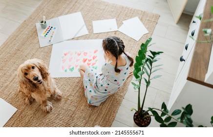 Ein Mädchen zieht Herzen für seine Mutter, die auf Teppichboden im Wohnzimmer sitzt, Cocker Spaniel Hund sitzt in der Nähe