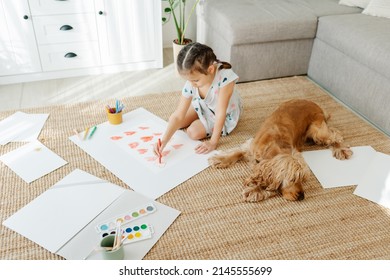 Ein Mädchen zieht Herzen für seine Mutter, die auf Teppichboden im Wohnzimmer sitzt, Cocker Spaniel Hund in der Nähe