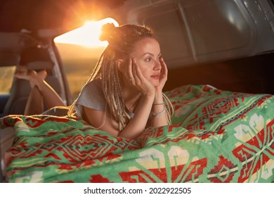 a girl with dreadlocks lies inside the car on a green blanket and looks away, the bright sun in the background. High quality photo