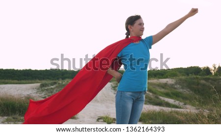 girl dreams of becoming a superhero. beautiful girl superhero standing on the field in a red cloak, cloak fluttering in the wind. Slow motion. young girl walks in red cloak expression of dreams