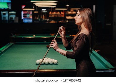 A girl in a dress stands with a cue in her hands and cleans it with chalk in a billiard club with balls in her hands.Playing billiards.