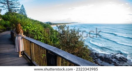 A girl in dress walks along the boardwalk and enjoys the view of the stunning Coolum Beach and surfers catching big waves, Pacific Ocean. Stunning panorama of Sunshine Coast, Queensland, Australia	