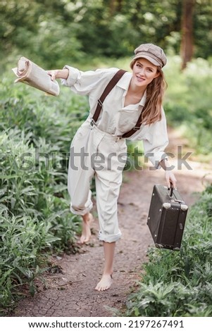 A girl dressed as a guy in retro style clothes. A girl dressed in retro style with a newspaper and a suitcase in her hands against the backdrop of nature. paperboy. crossdressing.