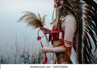 A girl dressed as a Native American with roach on her head holds herbs and a mascot in the hands standing by the river - Shutterstock ID 1659133606