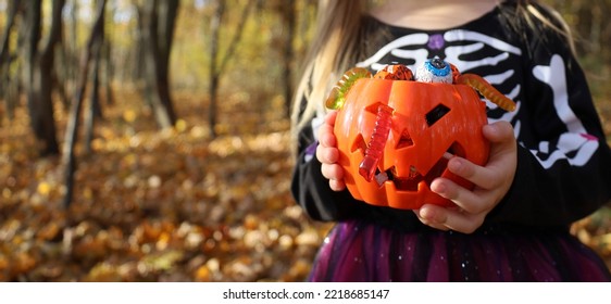 Girl Dressed Skeleton Costume Holds In Hands Orange Pumpkin Plastic Bucket Full Of Traditional Candies And Jelly Worms. Halloween Trick Or Treat Concept. Banner, No Face, Unrecognizable. Forest Behind