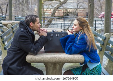 A Girl Dressing In A Blue Faux Fur Plus Size Jacket And  A Guy Dressing In A Black Pea Coat Are Sitting Crossing A Table, Looking At Each Other, With A Leather Bag. / Two Friends And One Bag 