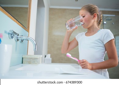 Girl drinking water while holding toothbrush by sink in bathroom at home - Powered by Shutterstock