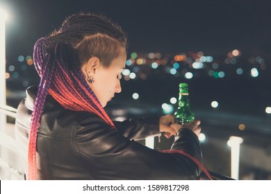 Girl Drinking Whiskey From A Bottle In An Attempt To Forget. Drunk Girl On The Background Of The City At Night On A High Floor Looking Down. Atmosphere Of Sadness And Sorrow