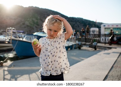 The girl is eating the aaple and scratching her head at the port be the sea at sunshine