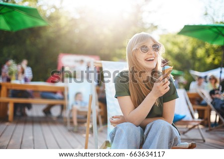 Girl eating ice cream laughing. Portrait of young female sitting in a park on a sunny day eating icecream looking off camera wearing glasses enjoying summer.