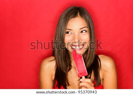 Girl eating popsicle ice pop looking cute to the side on red background. happy excited expression portrait on red background. Multicultural Caucasian / Chinese Asian young woman model.
